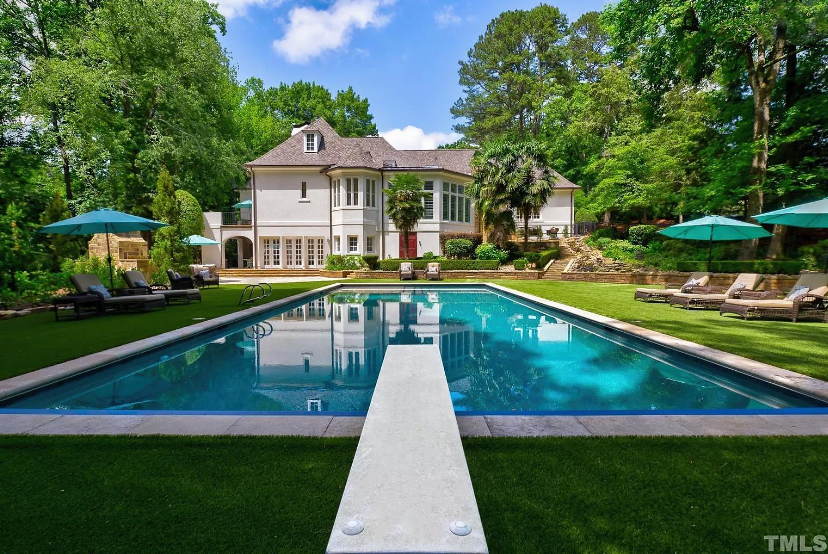 A lush, green backyard oasis with a clear, teal swimming pool, diving board, lounge chairs with umbrellas, and a large white house